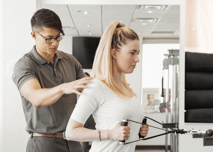 A practitioner guiding a patient through an exercise on a functional pulley machine, one hand on her shoulder blade.