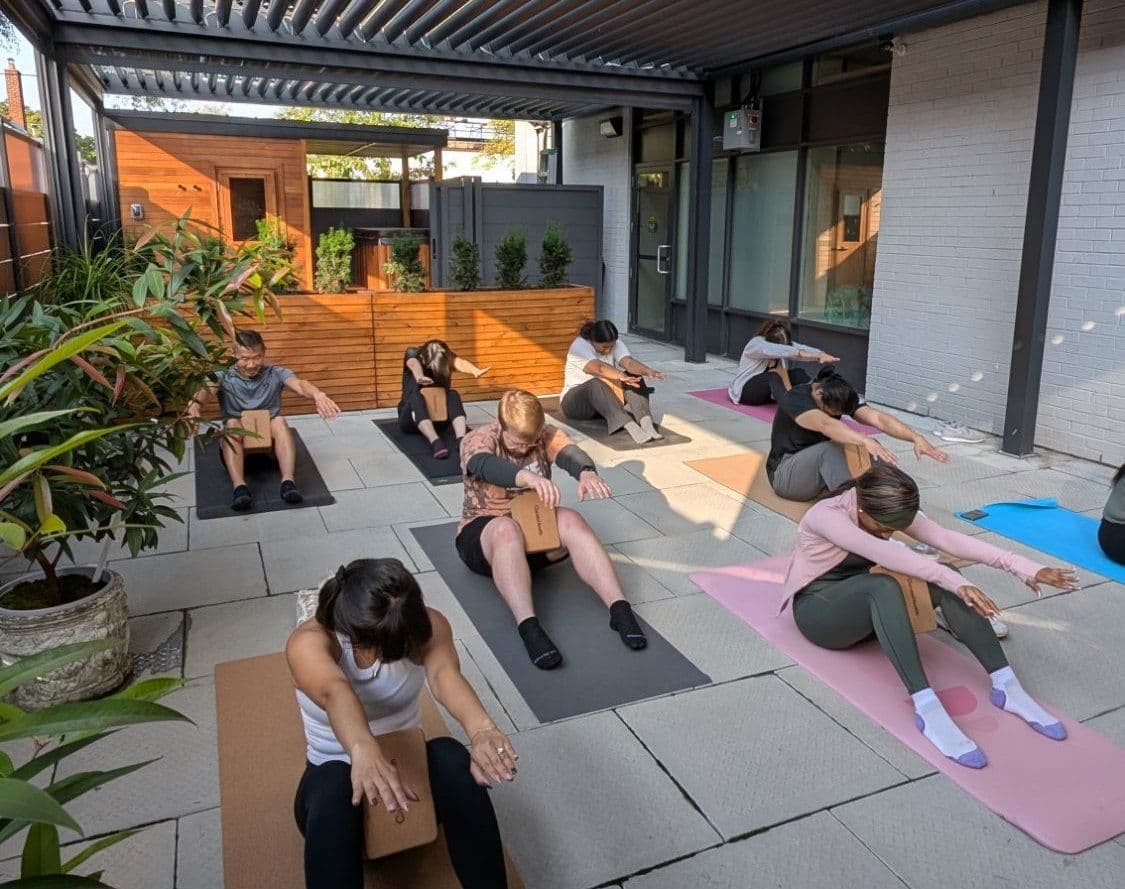 An Outdoor Pilates class seated on mats under the pergola, reaching to one side with cork blocks, with the sauna cabin and planted beds behind them.