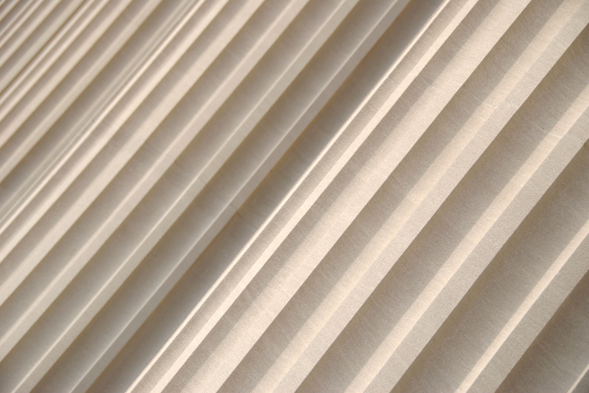 Close-up photograph of diagonal beige or cream-colored pleats creating a repeating linear pattern with light and shadow.