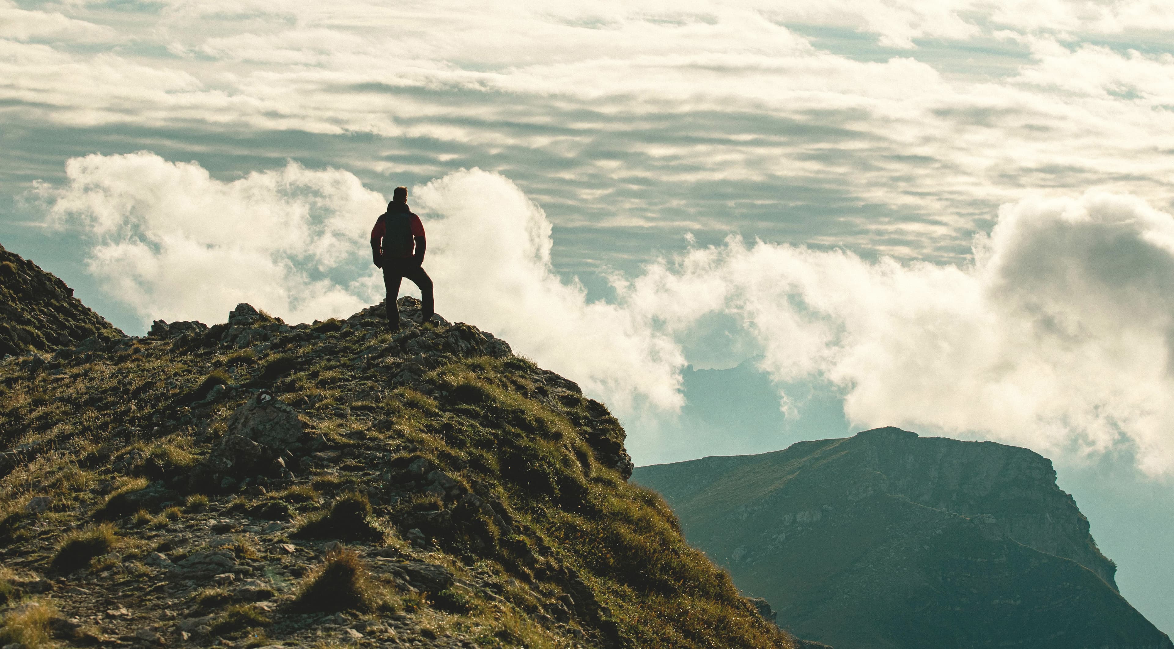 A silhouette on the top of a mountain in the foreground with clouds shown in the background.