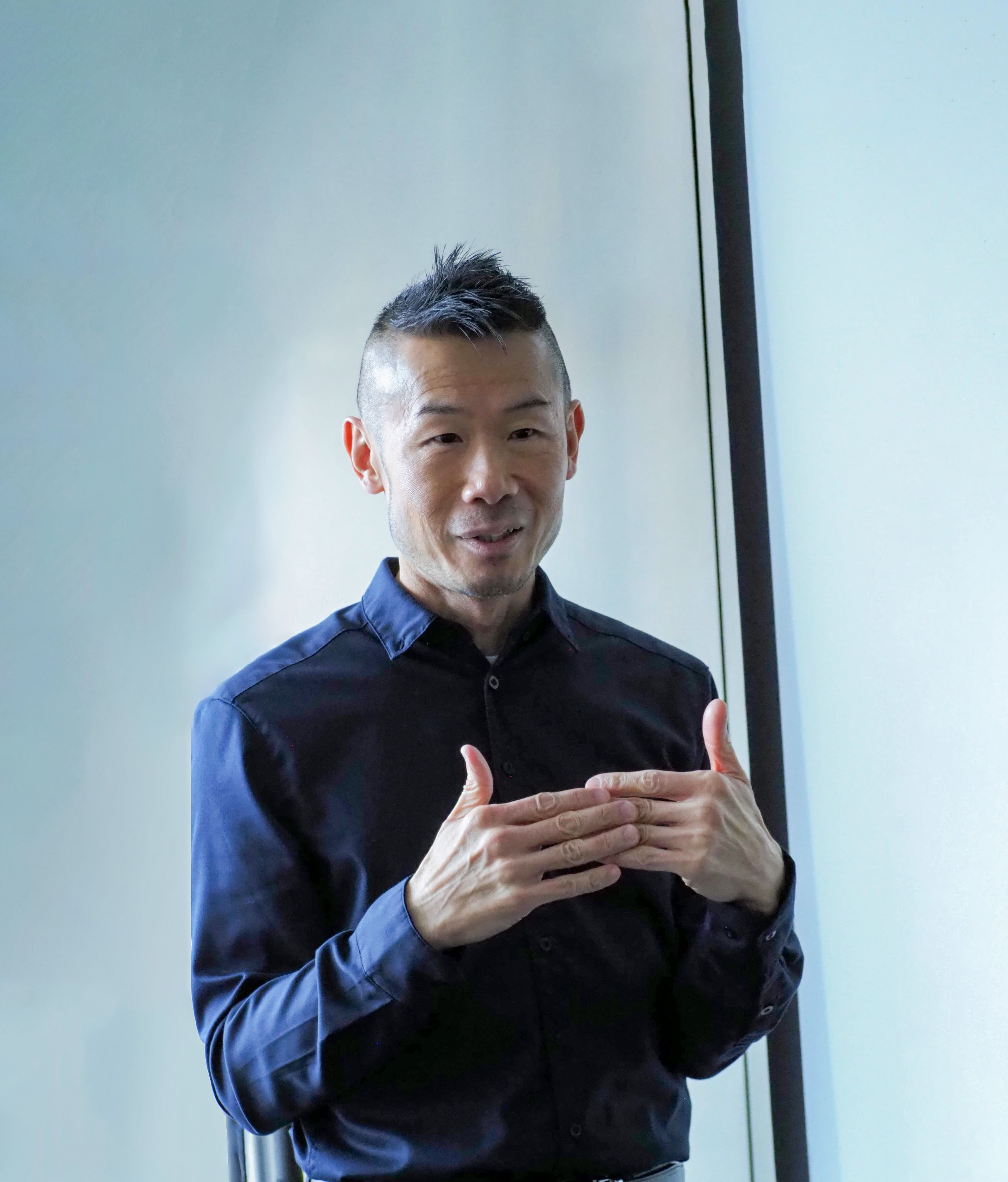 Dr. Eric Leong standing beside a window in a dark shirt with his hands clasped.