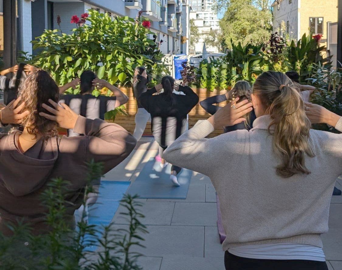 An Outdoor Pilates class under the pergola in the Recovery Sanctuary, hands behind their heads, facing the planted beds and the street beyond.