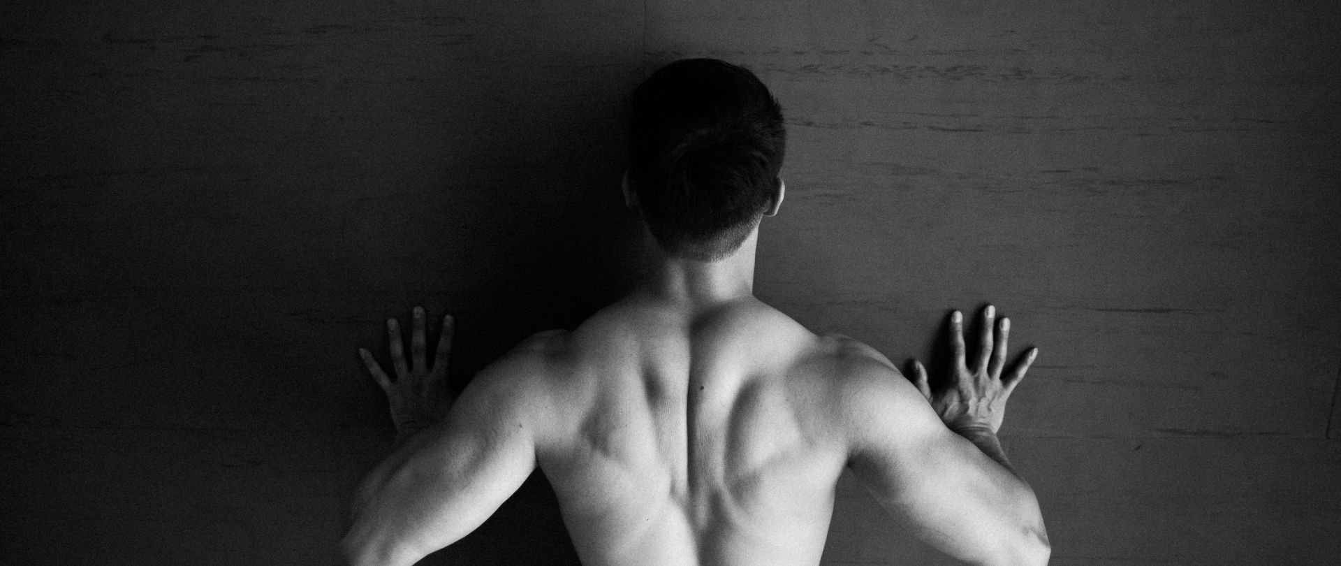 Black-and-white photo of a muscular man viewed from behind, performing a push-up against a textured wall.