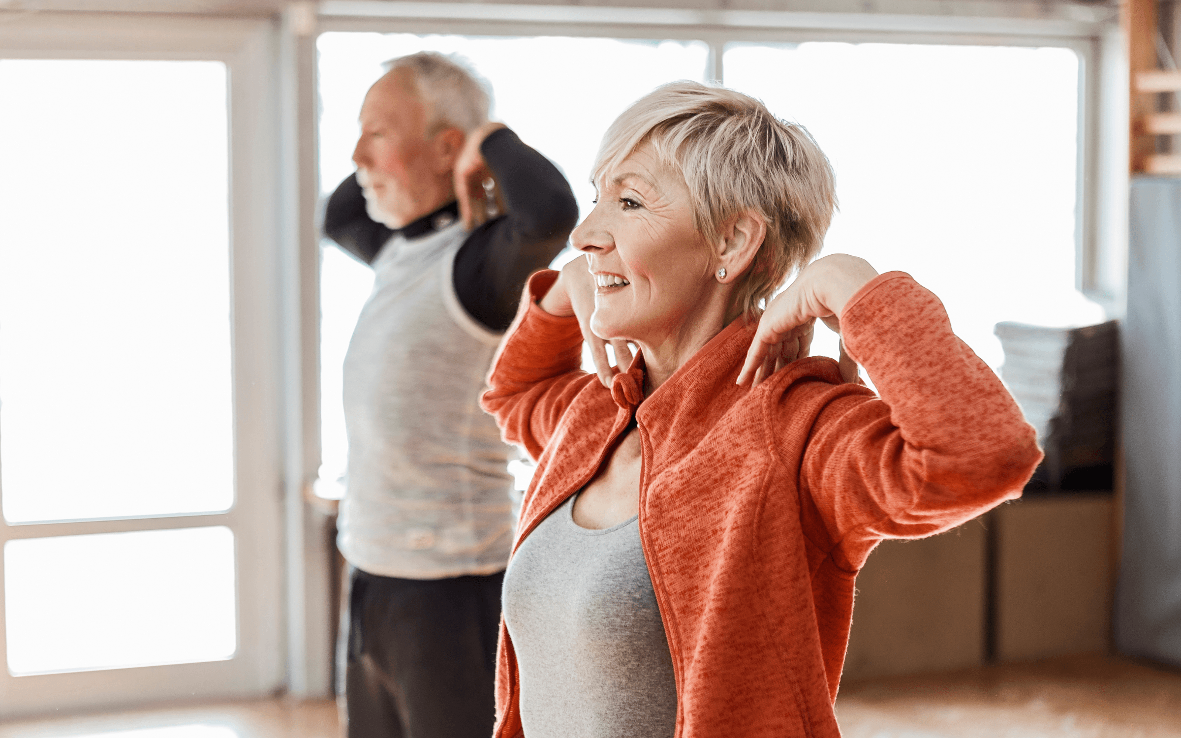 Smiling mature woman in athletic wear holding a kettlebell during a group fitness class, with other participants visible in the background