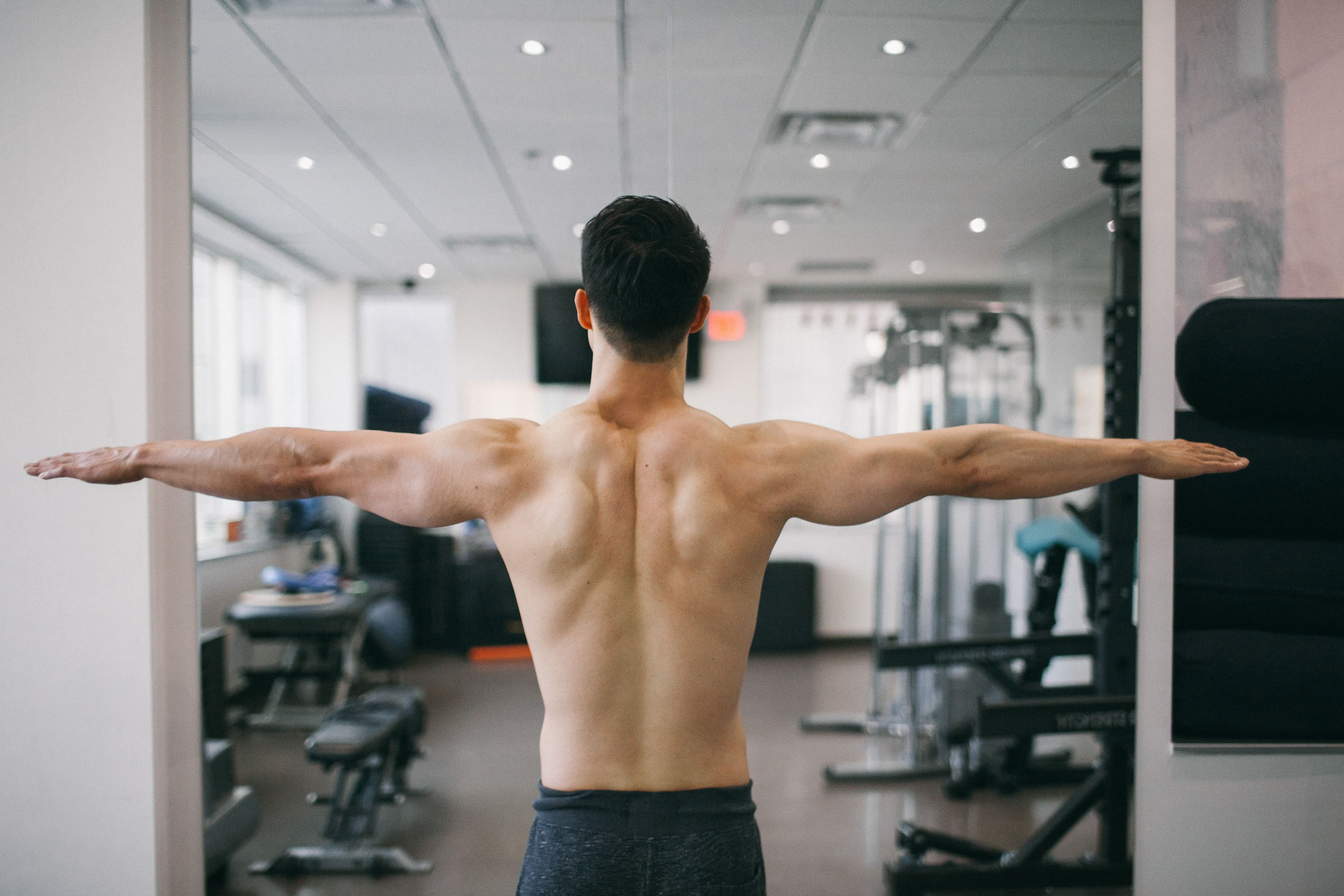 Rear view of man with a muscular back with open arms performing a shoulder mobility exercise in a fitness facility with exercise equipment visible in the background
