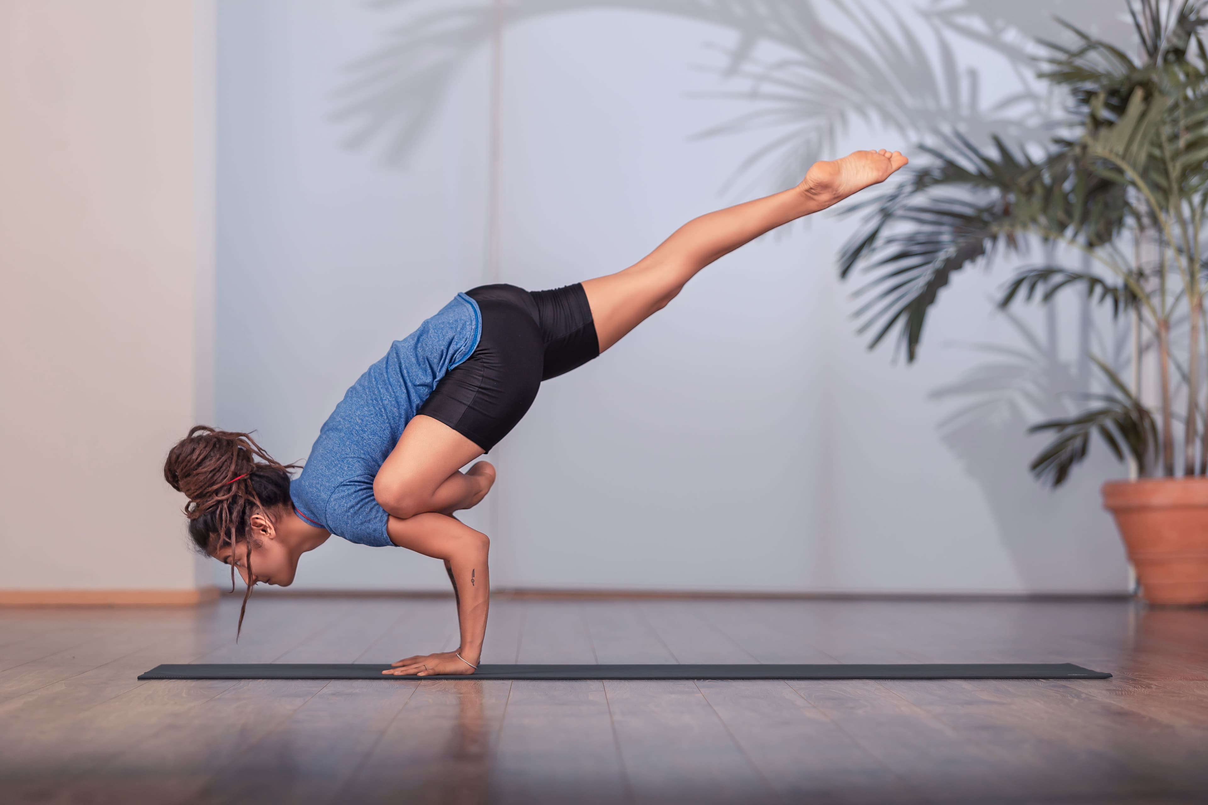 Rooj Hussain balancing in an arm balance yoga pose on a mat beside a potted palm.