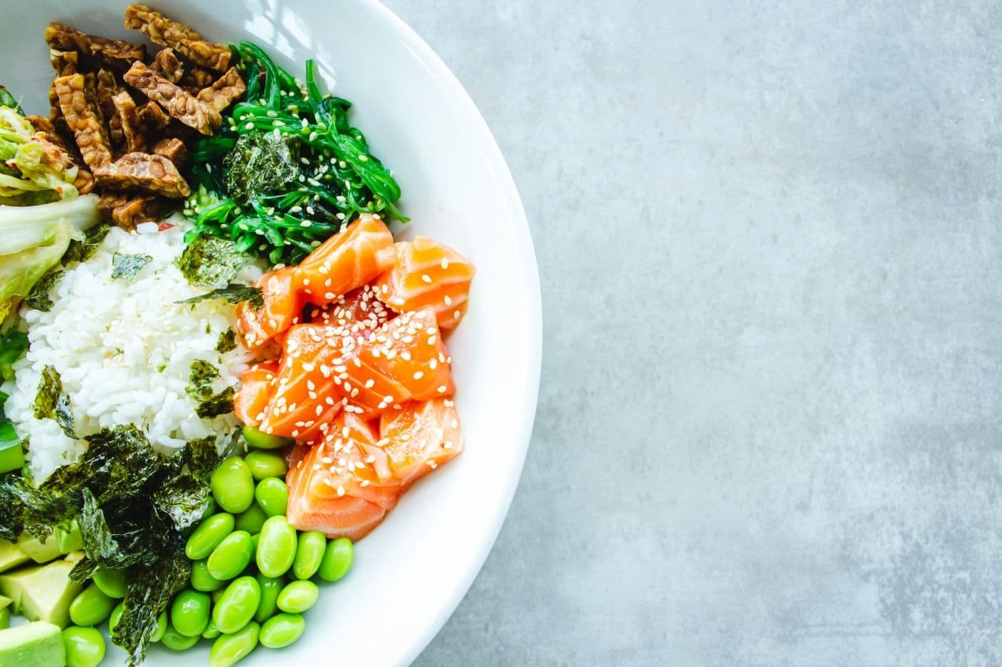 Overhead photograph of a poke bowl featuring fresh salmon sashimi, edamame, white rice, seaweed, and mixed vegetables garnished with sesame seeds.