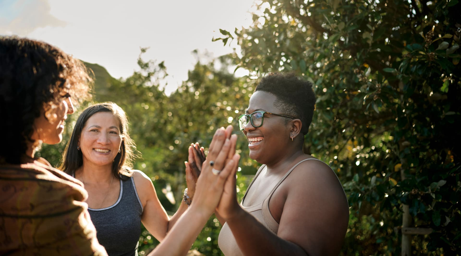 Photograph of three diverse women in athletic wear celebrating with high-fives outdoors during sunset or golden hour.