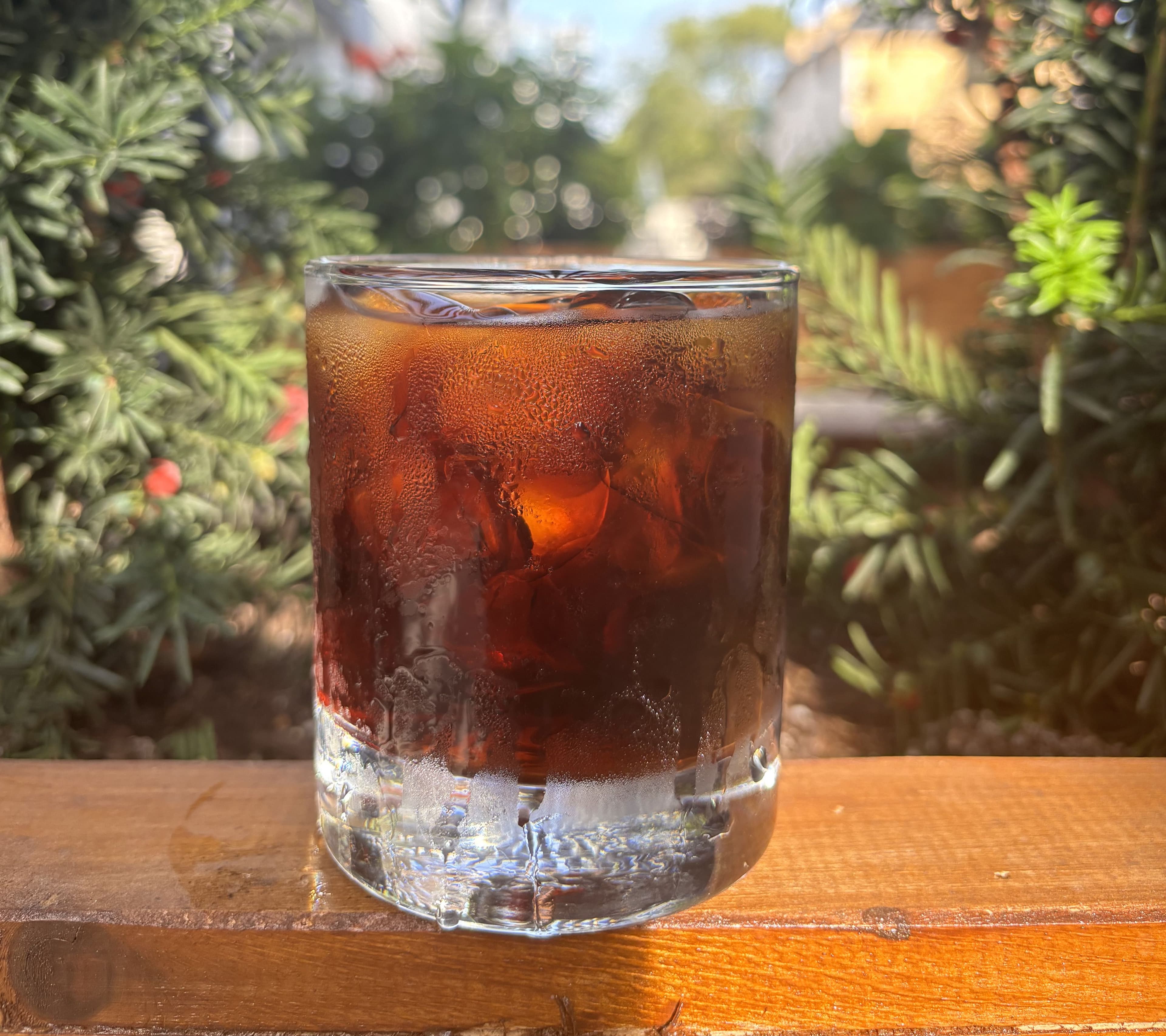 Iced coffee in a textured glass on a wooden rail, with greenery and a pergola behind.
