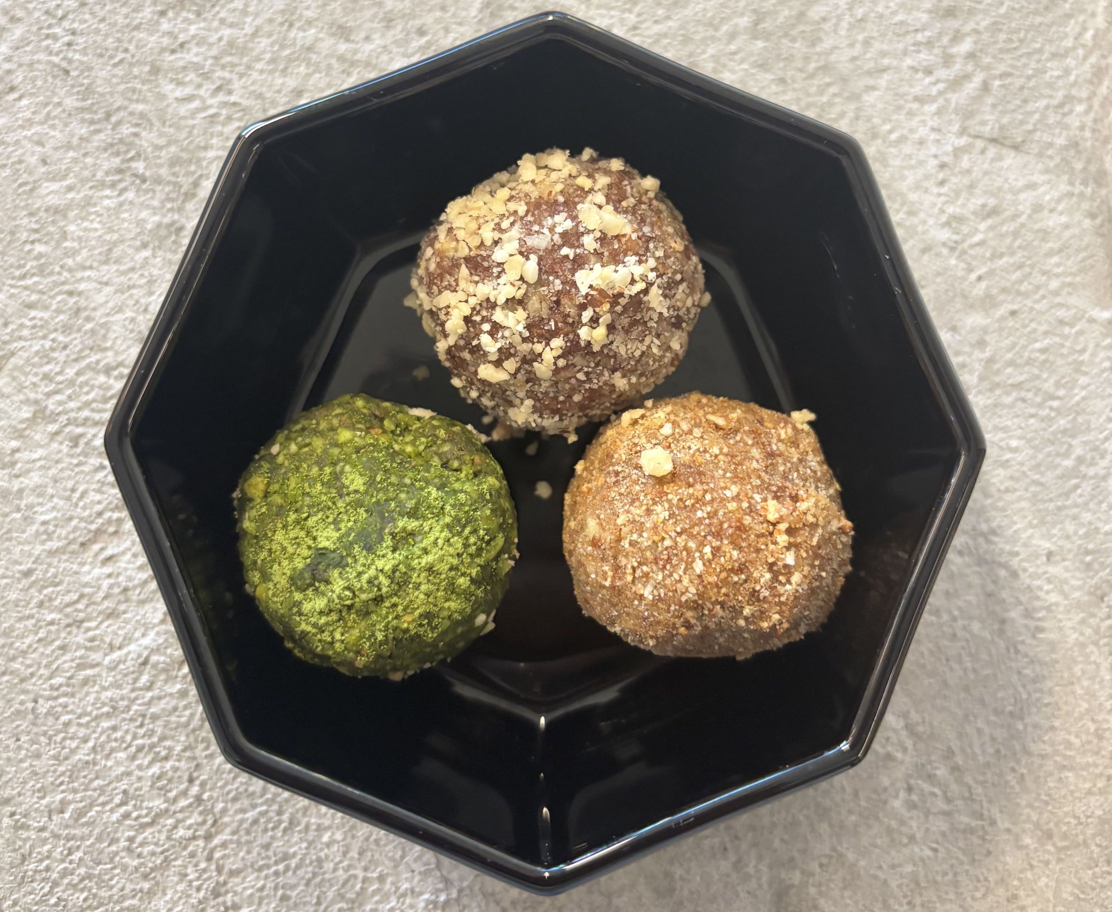 Three energy balls in a black bowl, rolled in matcha, chopped nuts and golden crumb.