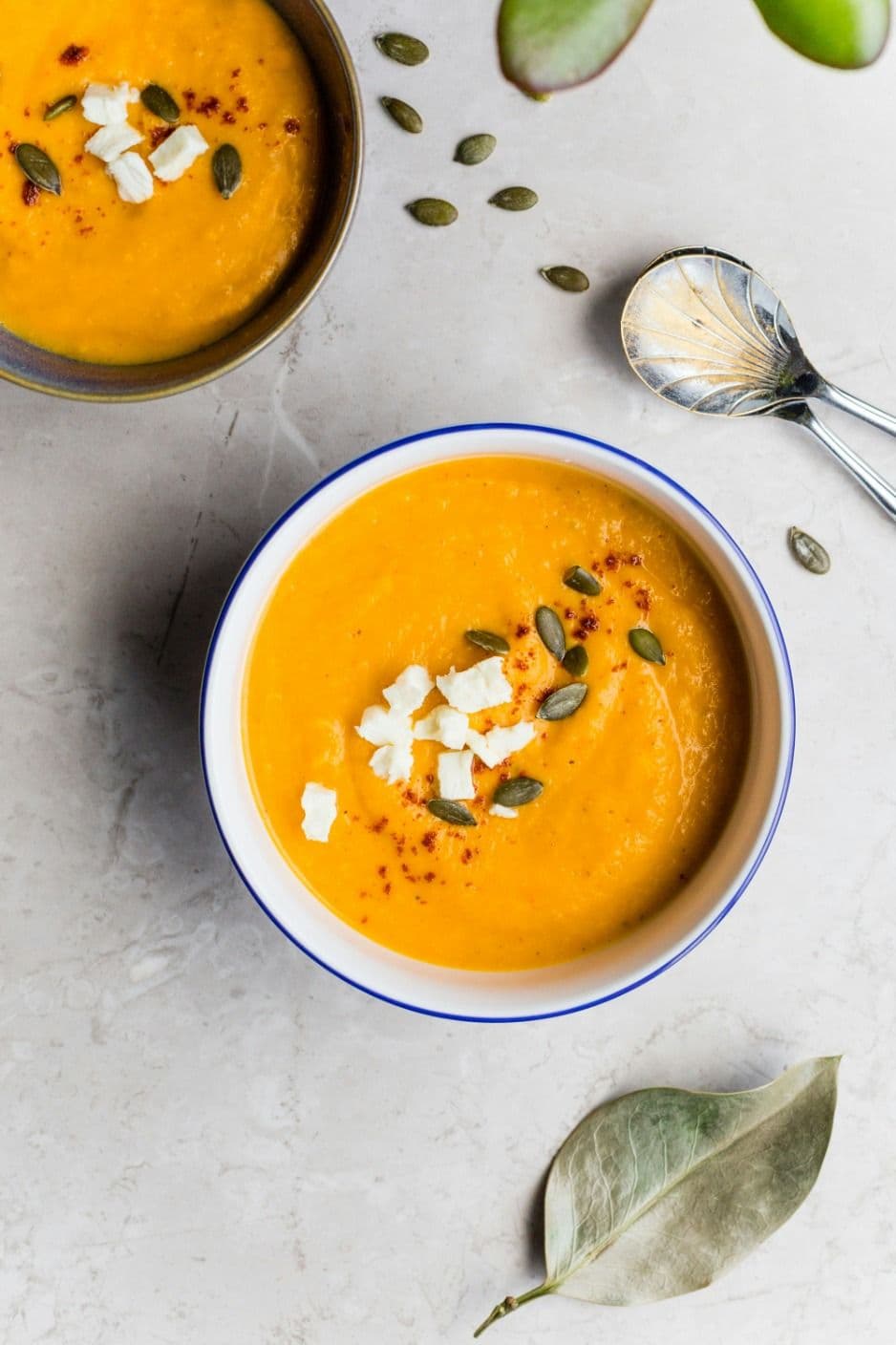 Overhead photograph of butternut squash soup served in white bowls with blue rims, garnished with crumbled cheese, pumpkin seeds, and spices on a light marble surface.