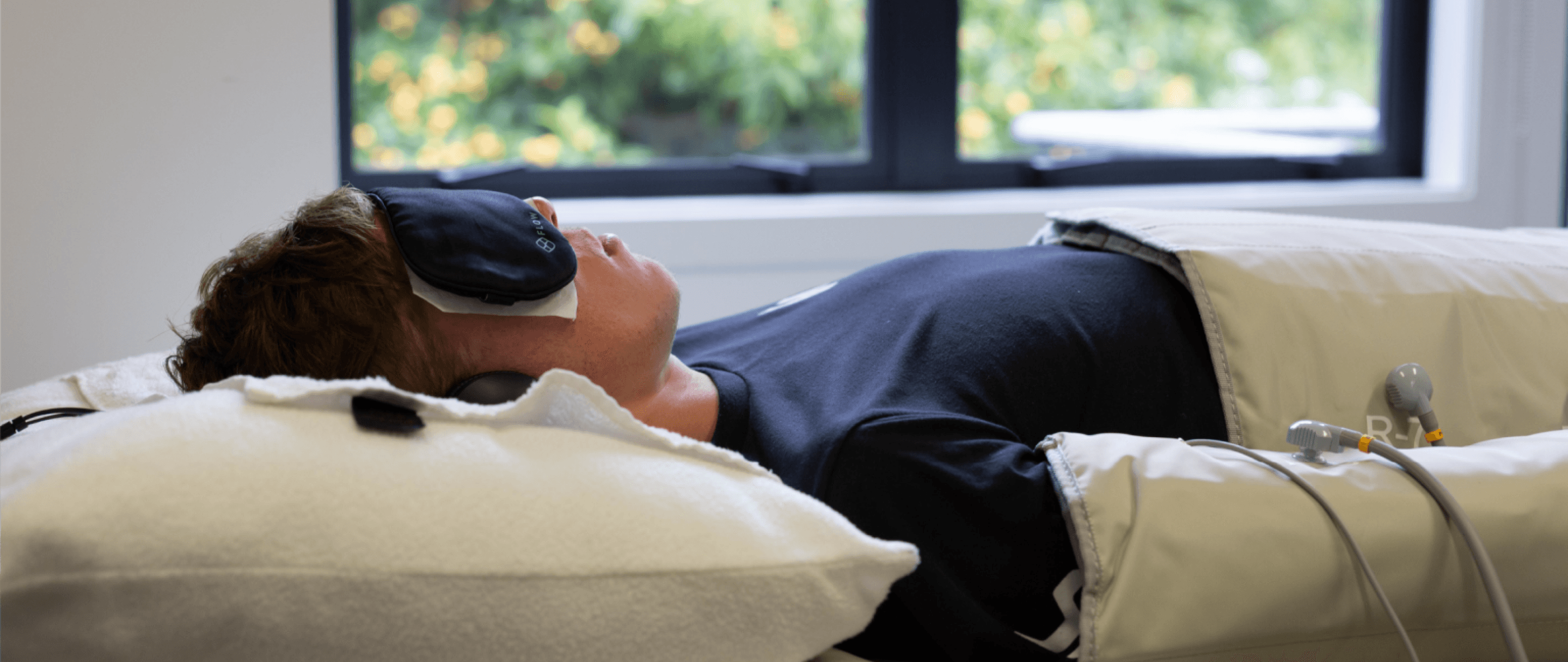 Photo of a person lying on a treatment bed in the flowpresso suit with an eye mask feeling relaxed.