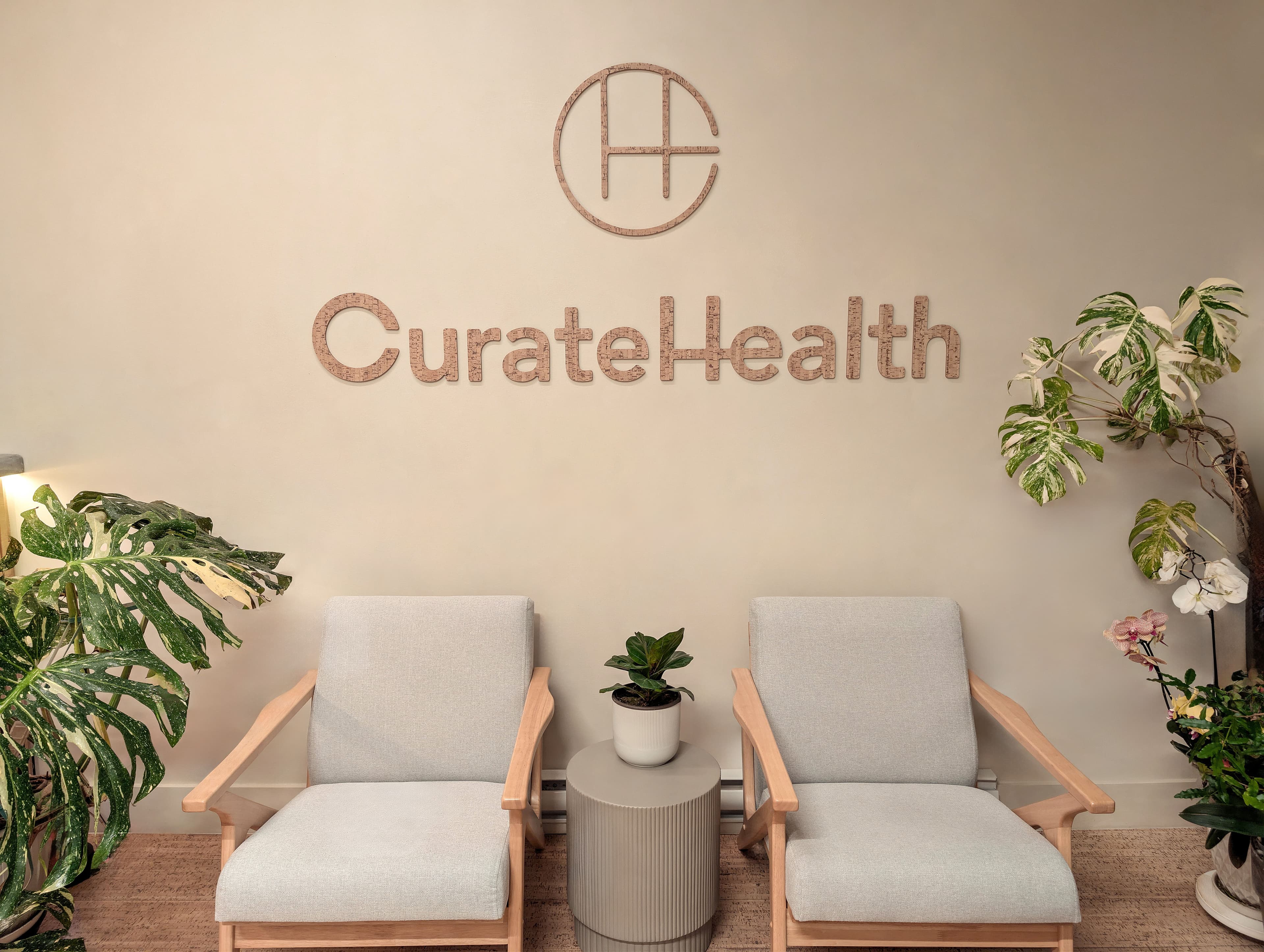 Two light wood armchairs below the Curate Health logo in cork lettering on a plaster wall, with plants on either side.