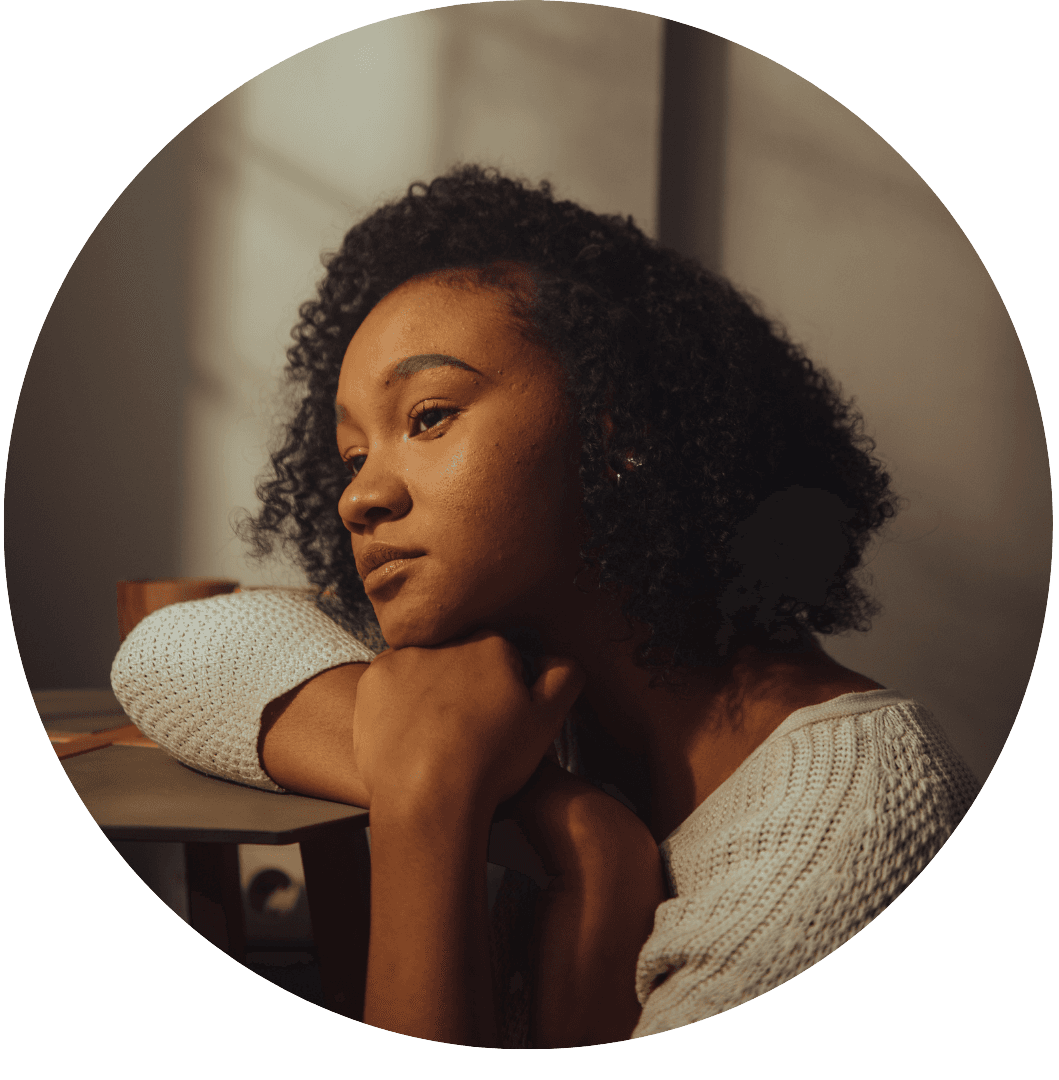 Portrait of a young Black woman in a white sweater resting her chin on her hand, with a thoughtful expression in warm lighting, cropped in a circular frame"